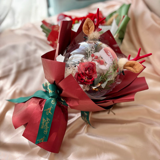 a delightful combination of a Red Rose and a Santa Claus Doll, enclosed in a crystal sphere with deer horns. Wrapped in elegant red paper, this preserved bouquet is a captivating and charming gift.